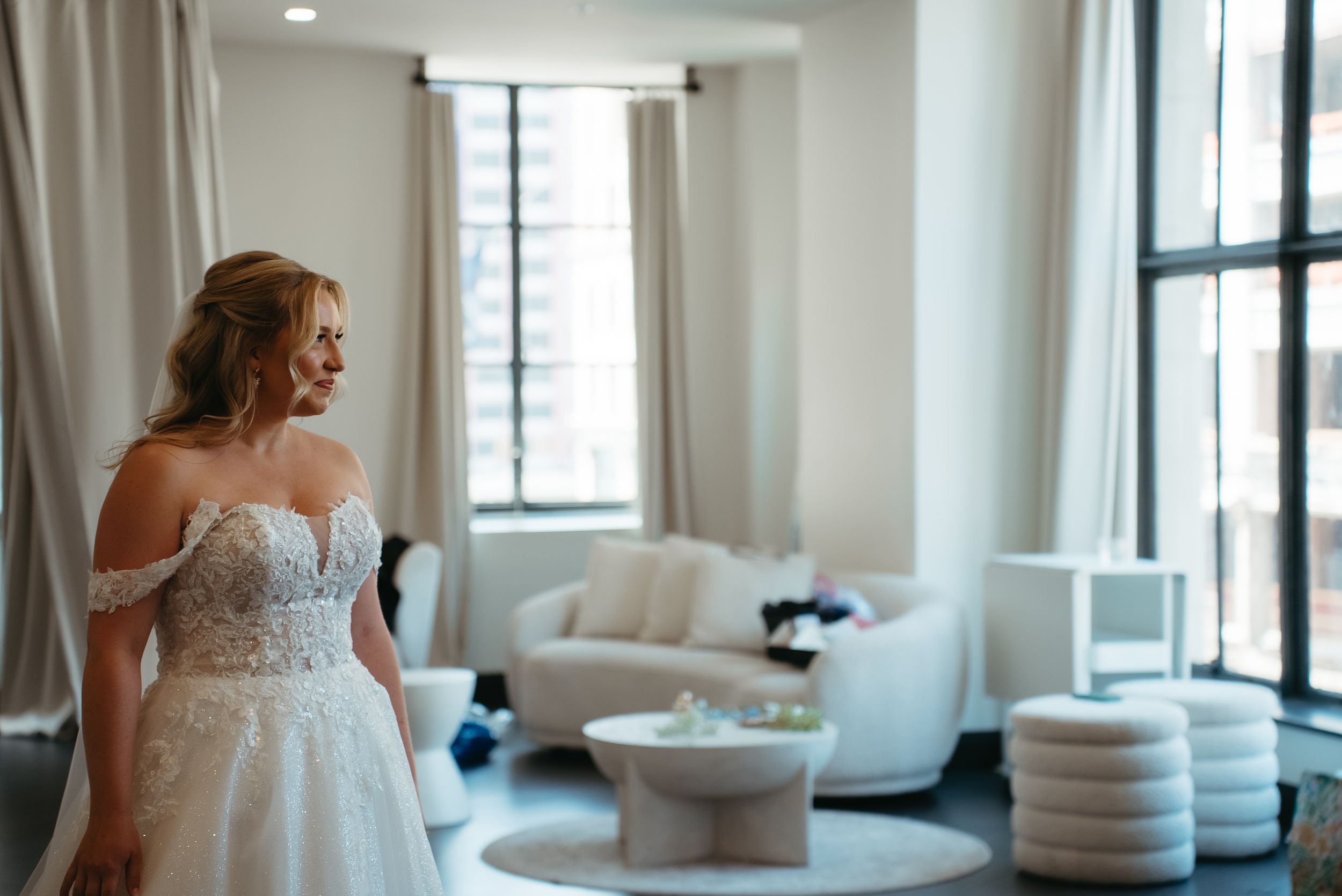 Bride in an off-shoulder lace ball gown wedding dress standing in a bright modern bridal suite.