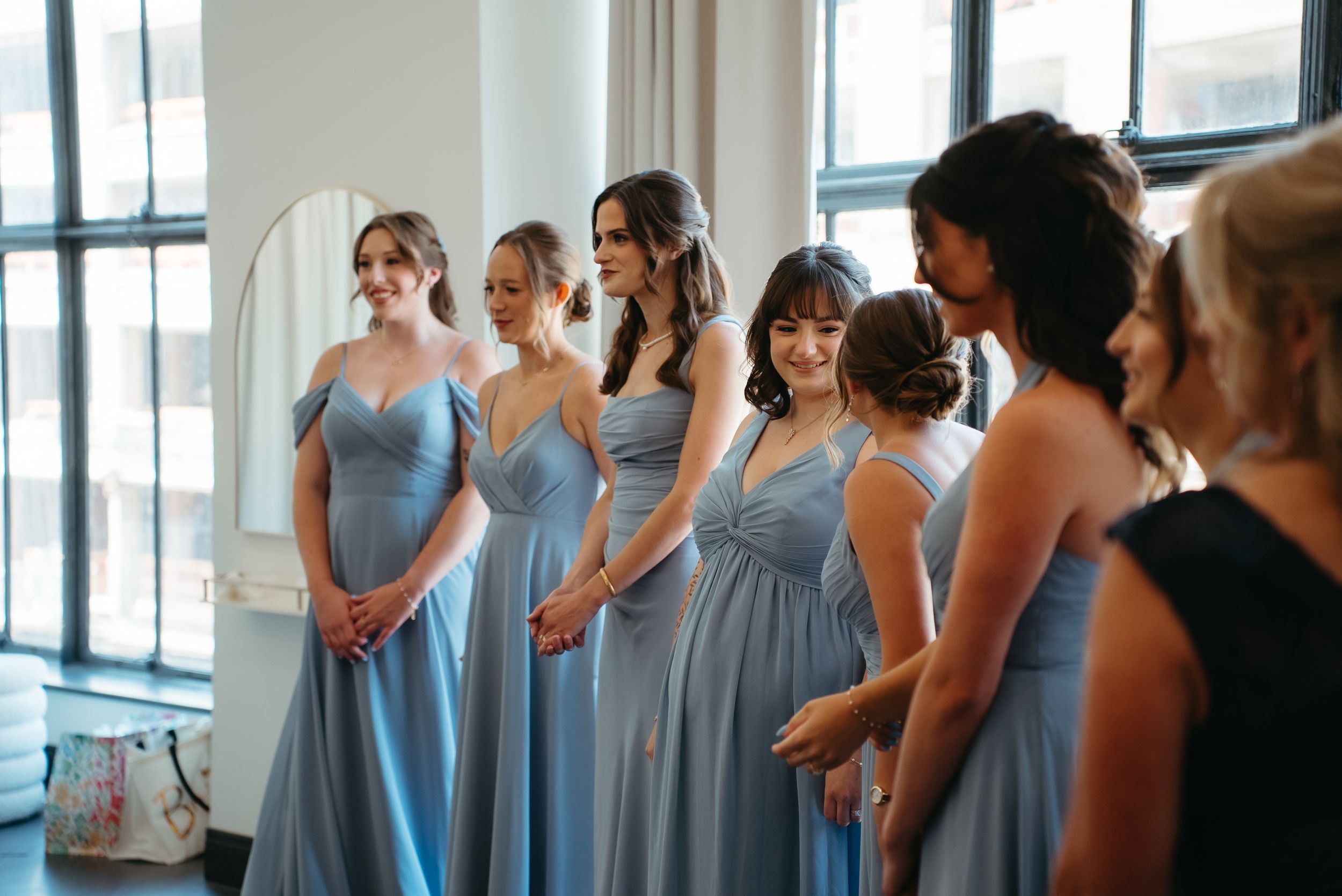 Bridesmaids in matching dusty blue gowns stand in a row during wedding preparations in a bright modern venue.