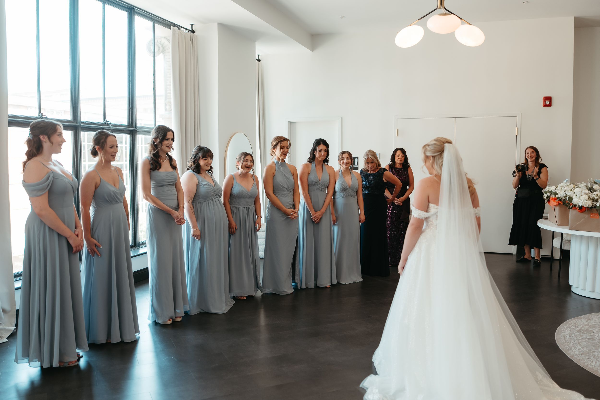 Bride in white gown and veil faces her bridesmaids in matching light blue dresses during a first look reveal.