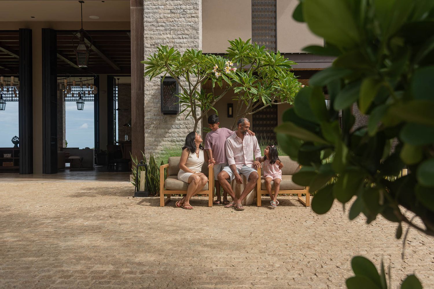 Family relaxing together on outdoor wooden furniture at a luxury tropical resort surrounded by lush greenery.