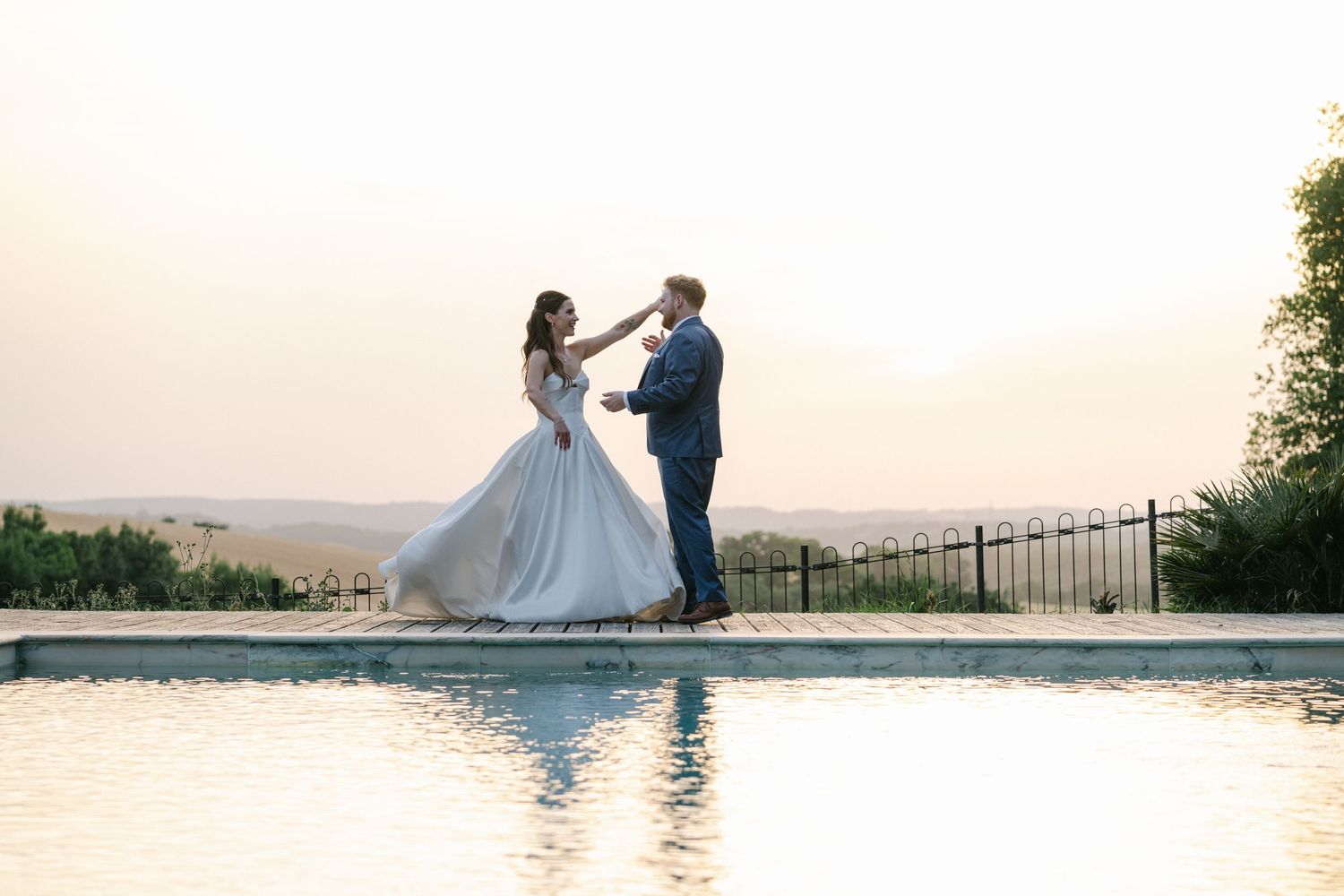 Bride and groom dancing by a pool at sunset with scenic hills in the background.