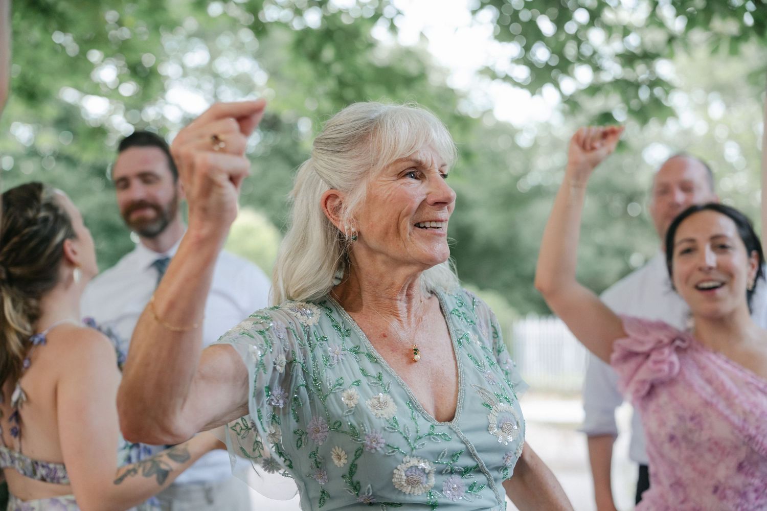 Elderly woman in floral dress dancing joyfully at an outdoor wedding celebration with guests in background.