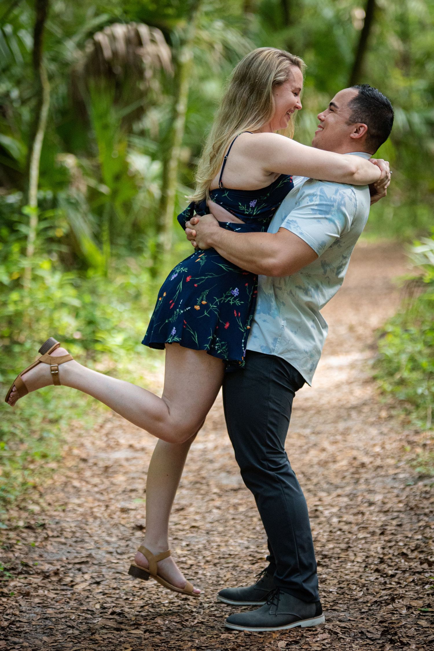 A joyful couple embraces on a forest path, the man lifting the woman in a floral dress among lush green trees.