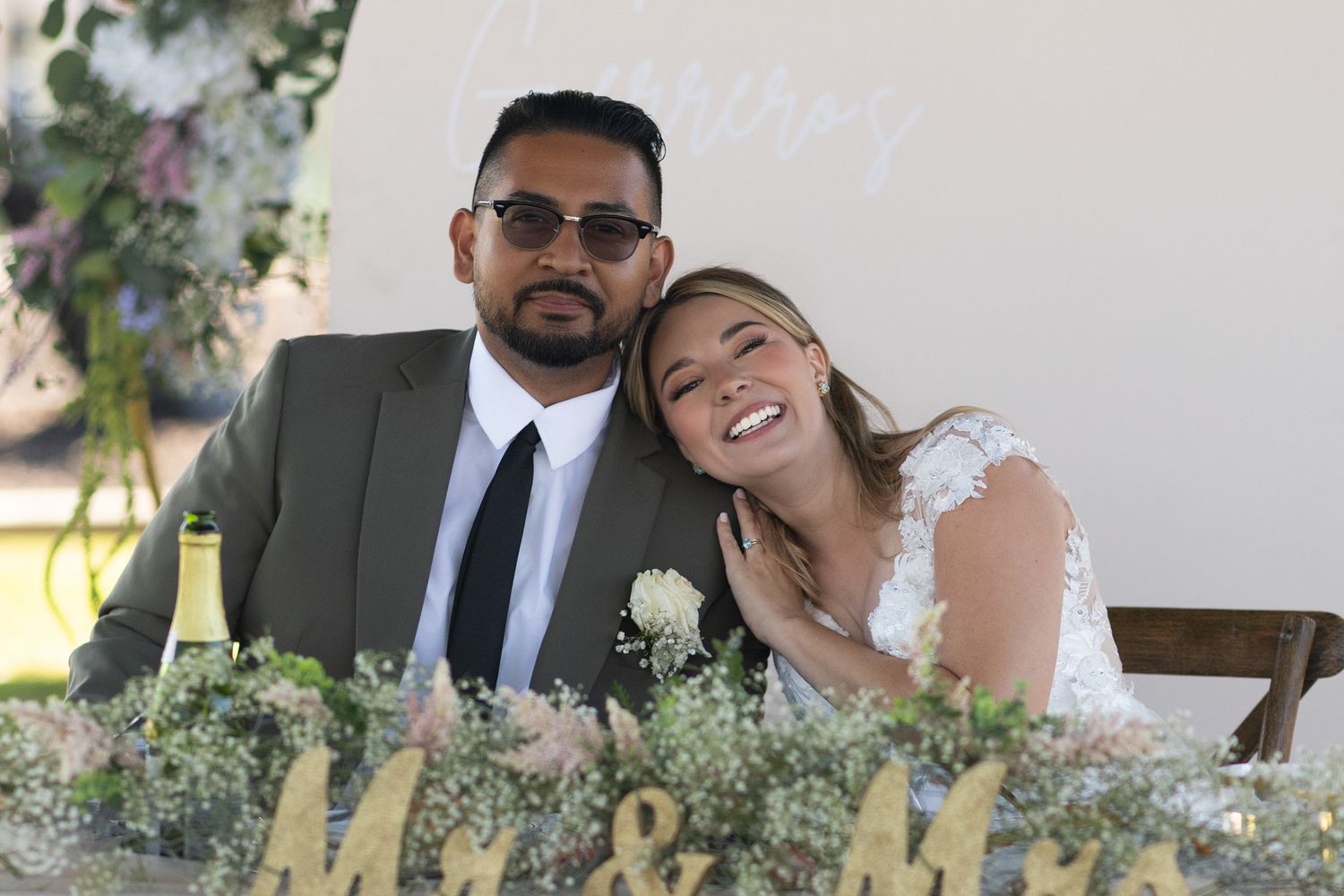 Happy couple poses at wedding reception table decorated with flowers and a Mr. & Mrs. sign.