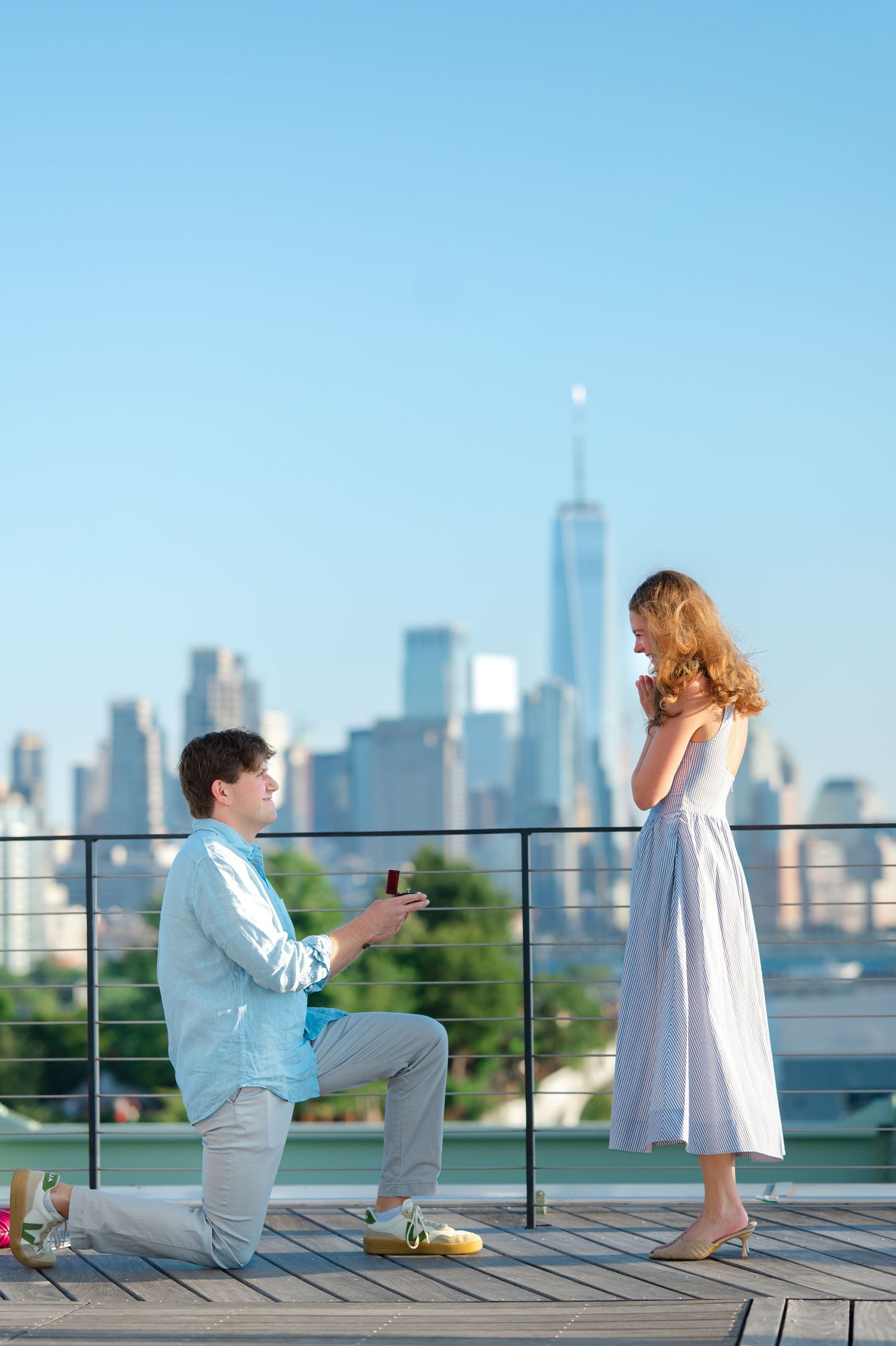 Man proposes to woman on rooftop terrace of Pier 57 in Chelsea with NYC skyline and One World Trade Center in background at sunset.