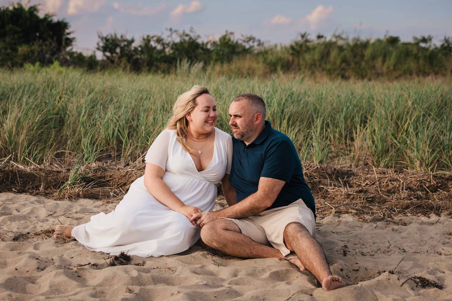 Engaged couple sitting together in tall dune grass at Duxbury Beach, Massachusetts, smiling in late afternoon light.