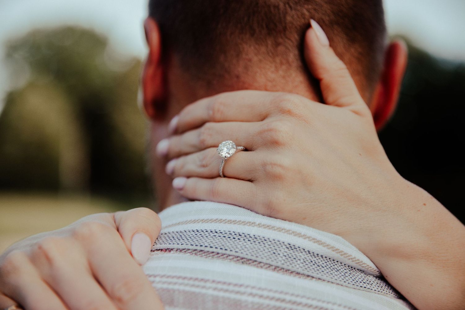 A woman\u2019s hand with a diamond engagement ring rests on a man\u2019s neck during a romantic outdoor moment.