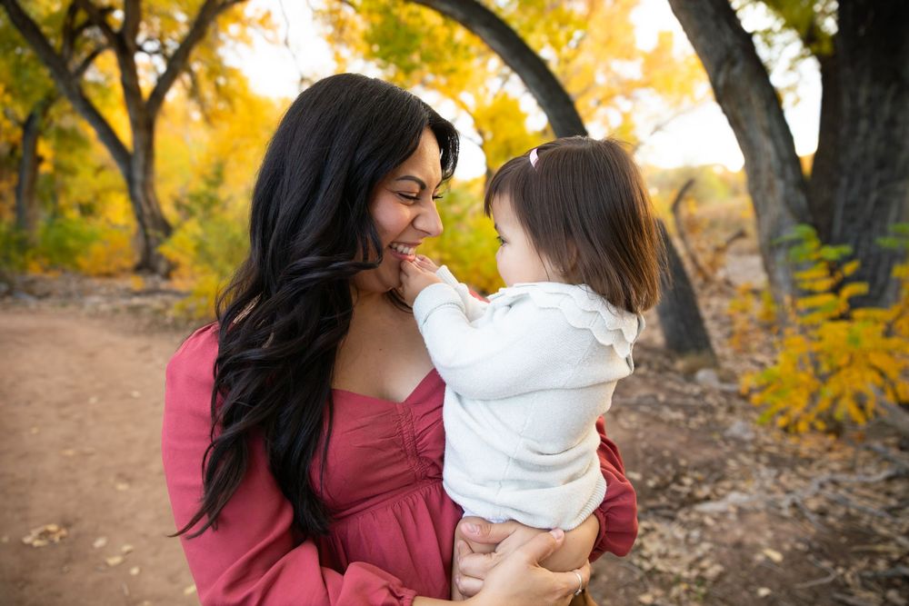 A mother in a red dress holds her toddler among autumn trees with golden leaves in a warm outdoor portrait.