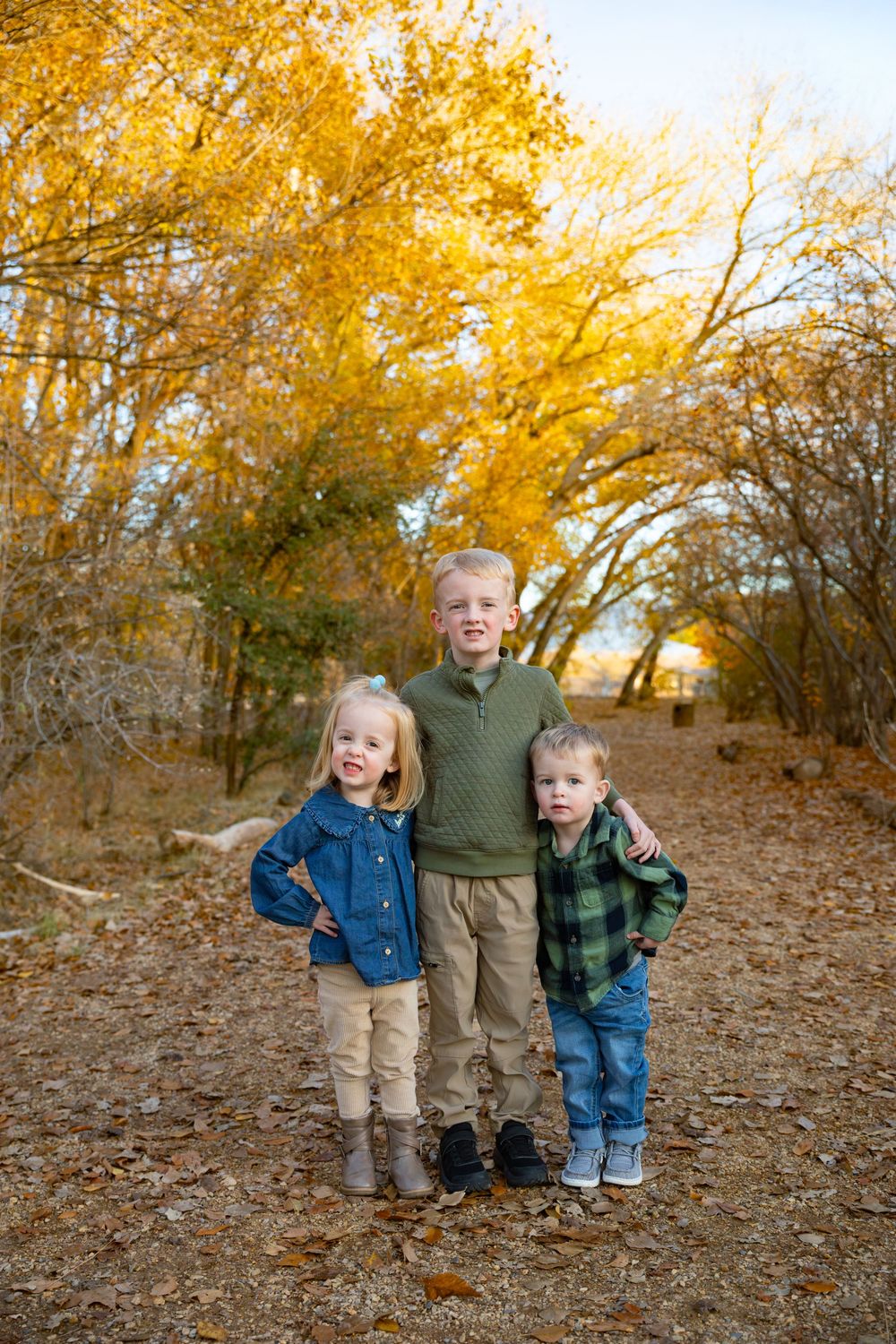 Three young siblings pose together on a leaf-covered path surrounded by vibrant golden autumn trees.