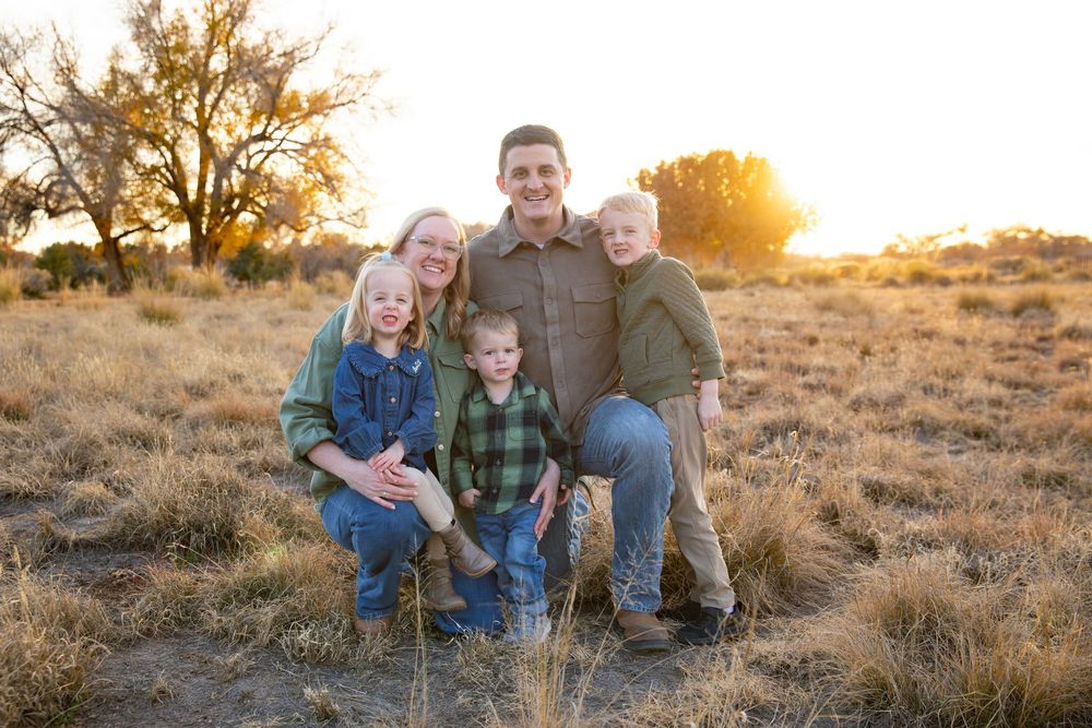 Family of five smiling outdoors in golden field at sunset, parents with three young children in casual attire.