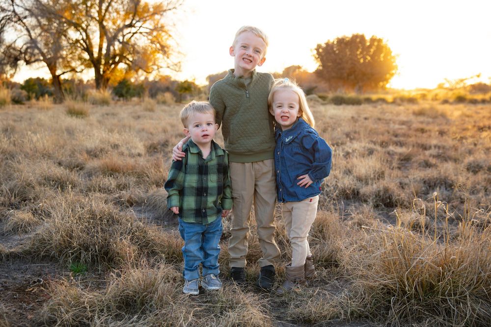 Three young children smiling together in a golden field at sunset, wearing casual outdoor clothing.