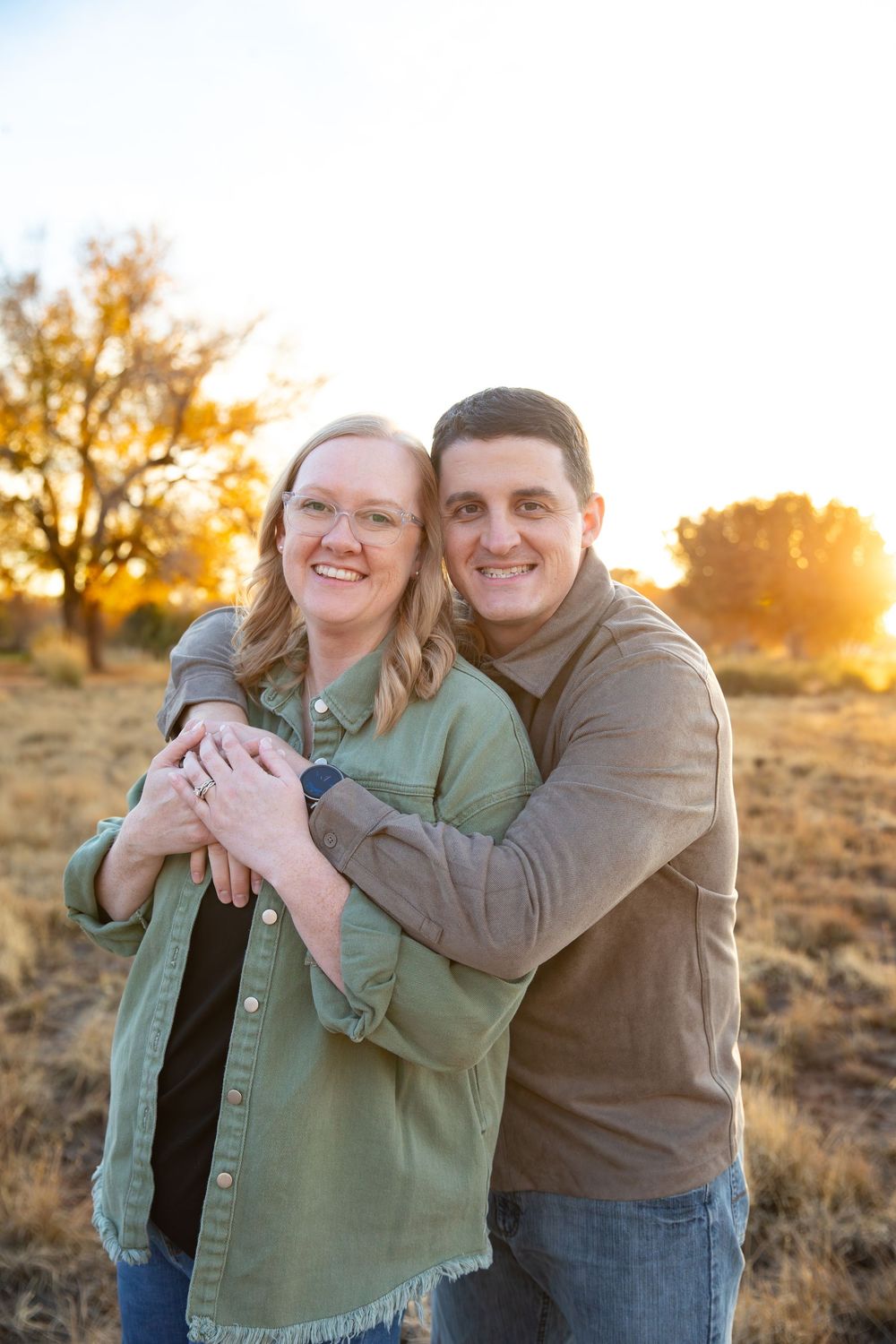 Couple embracing outdoors at golden hour, smiling warmly in a sunlit field with autumn trees in the background.