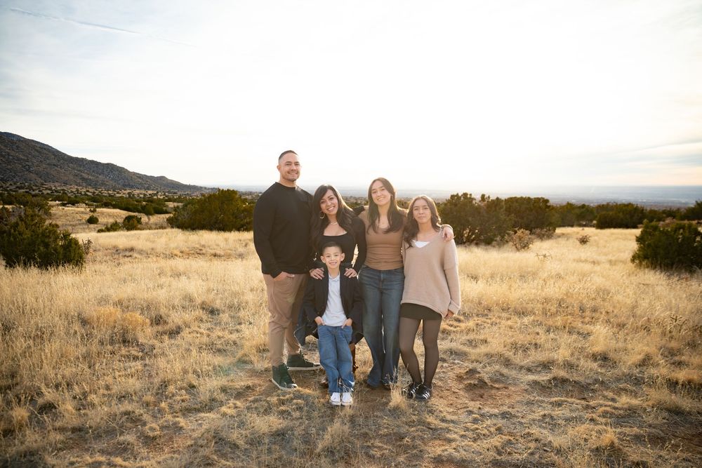 Family of five posing outdoors in golden meadow with mountain backdrop at sunset.