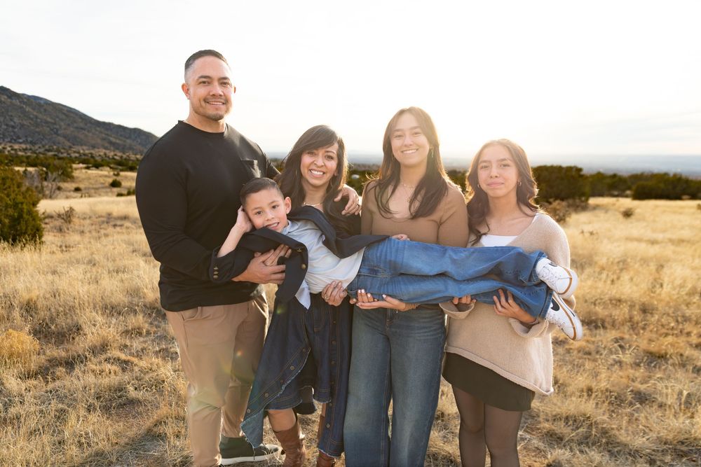 Family of five posing outdoors in a golden field with mountains at sunset, holding a baby wrapped in a blue blanket.