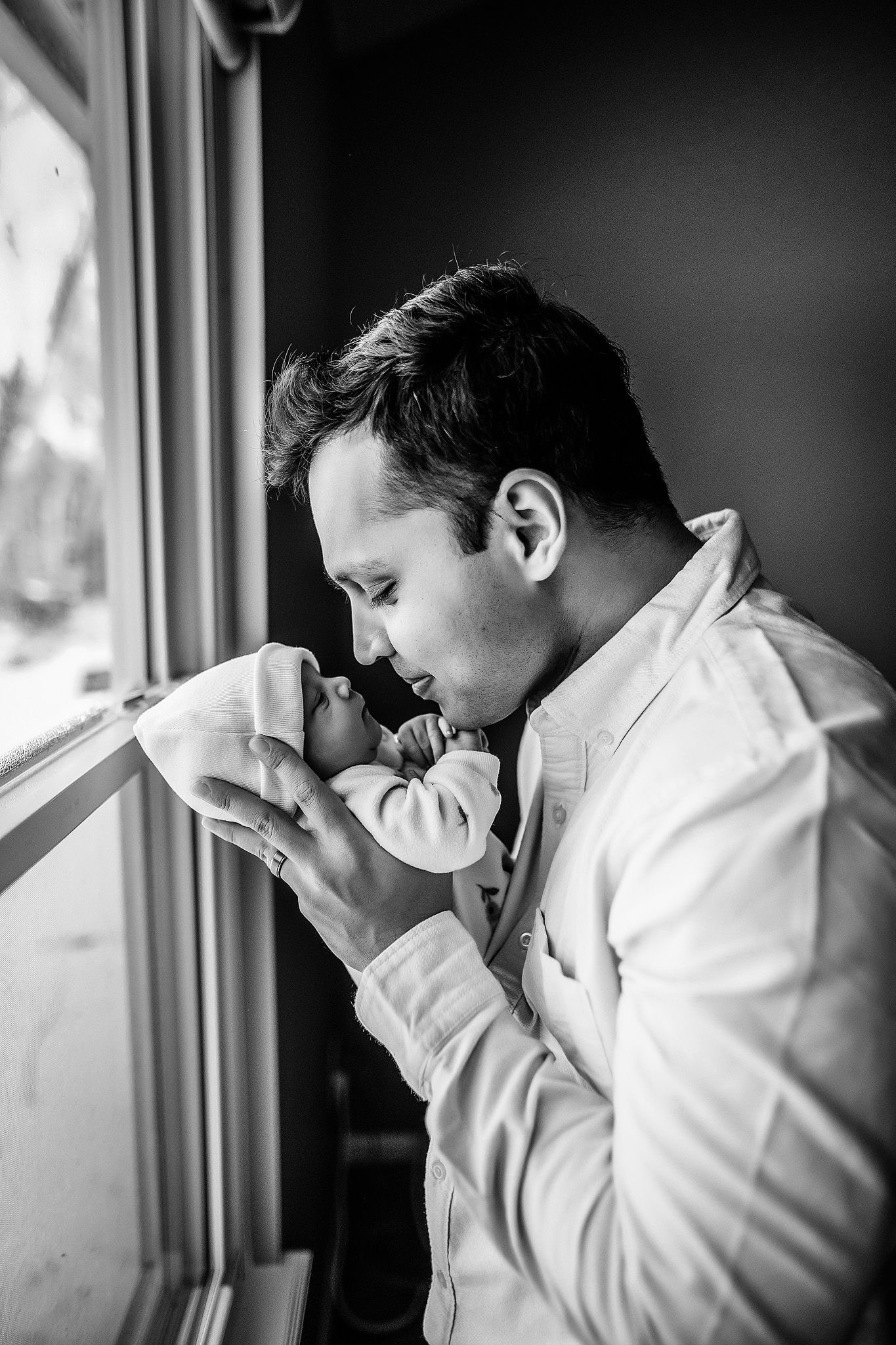 Black and white photo of a father tenderly kissing his newborn baby by a window.