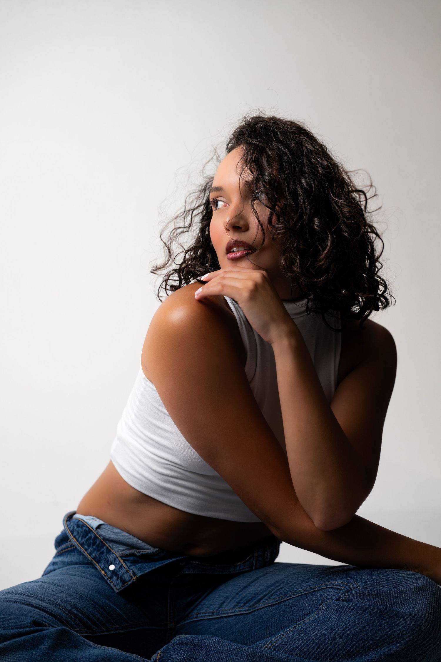 Young woman with curly hair in white crop top and jeans sits pensively against a light gray background.