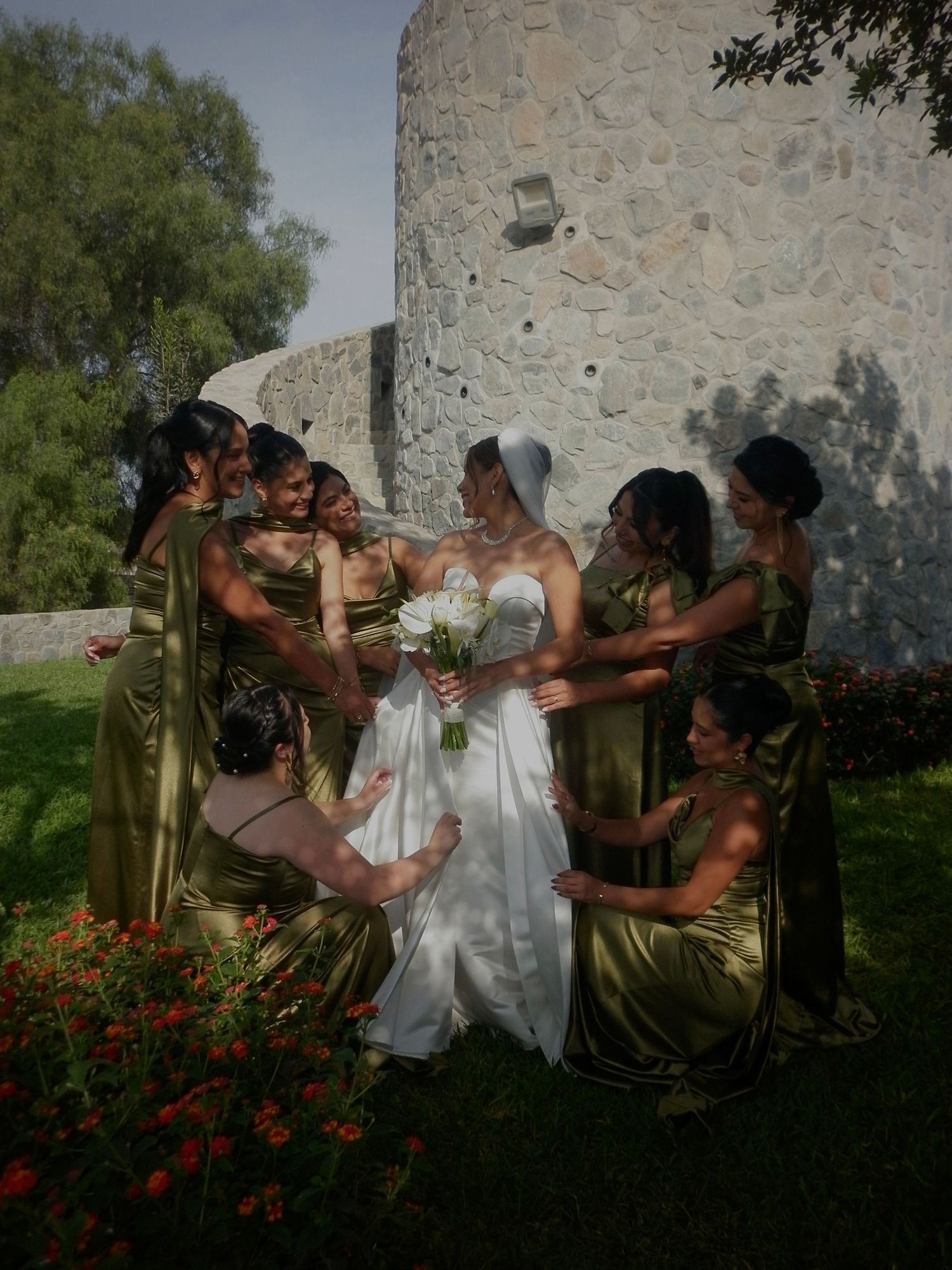 Bride in white gown with bridesmaids in gold dresses posing near stone tower surrounded by red flowers.
