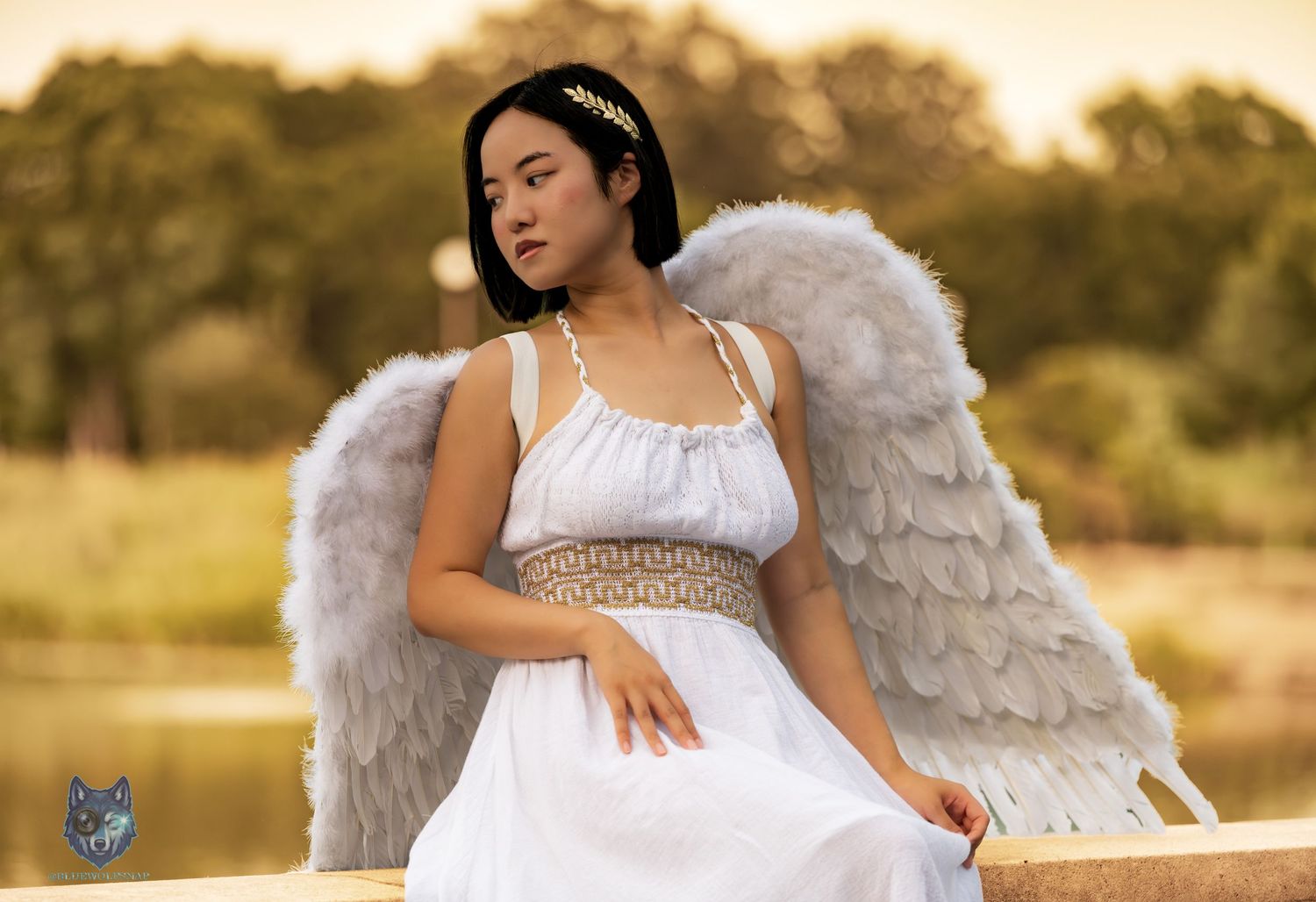 Young woman in white angel costume with feathered wings and gold crown sits outdoors near water.