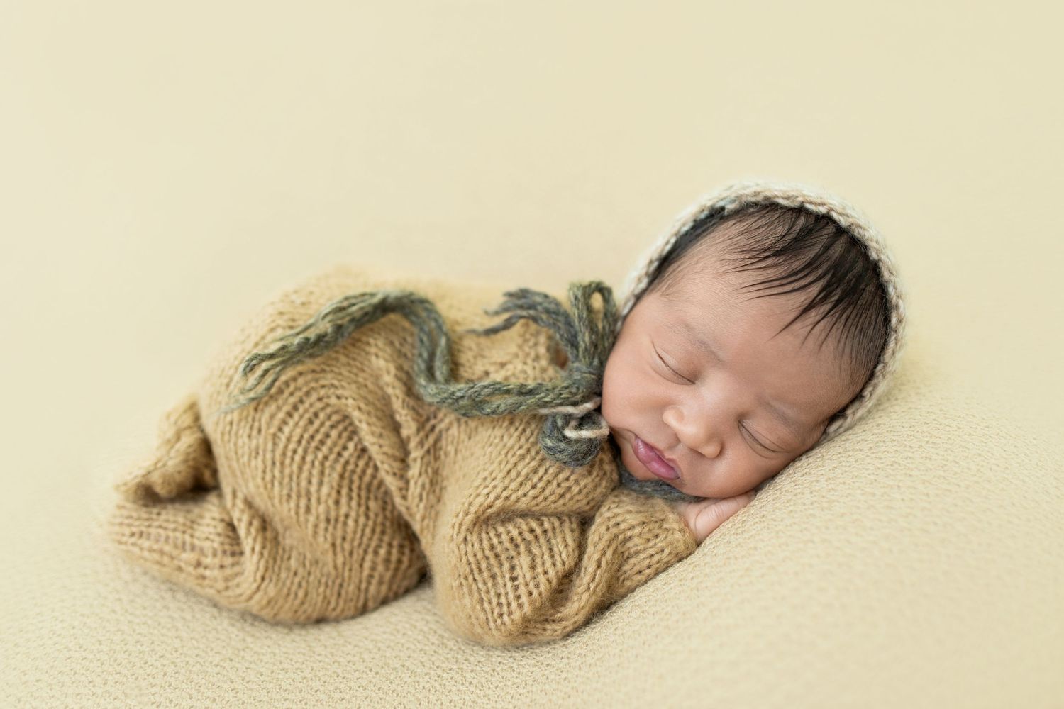 Sleeping newborn baby wrapped in a knit tan swaddle with lace bonnet on a soft yellow background.
