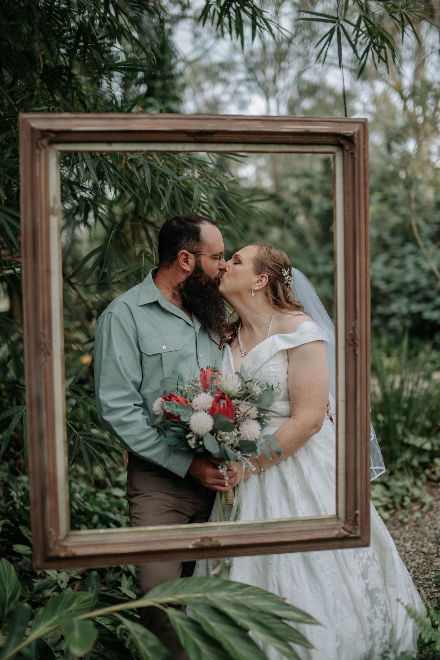 Bride and groom kissing inside a vintage picture frame surrounded by lush tropical greenery on their wedding day.