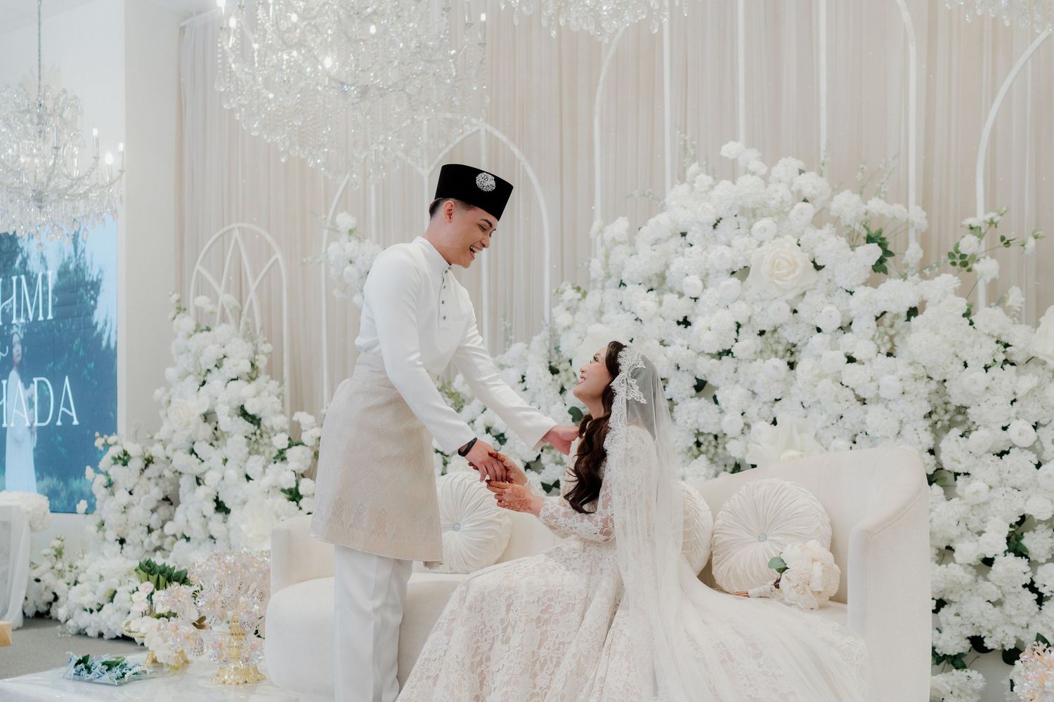 Elegant all-white Malay wedding ceremony with couple in traditional attire surrounded by lush white floral arrangements.