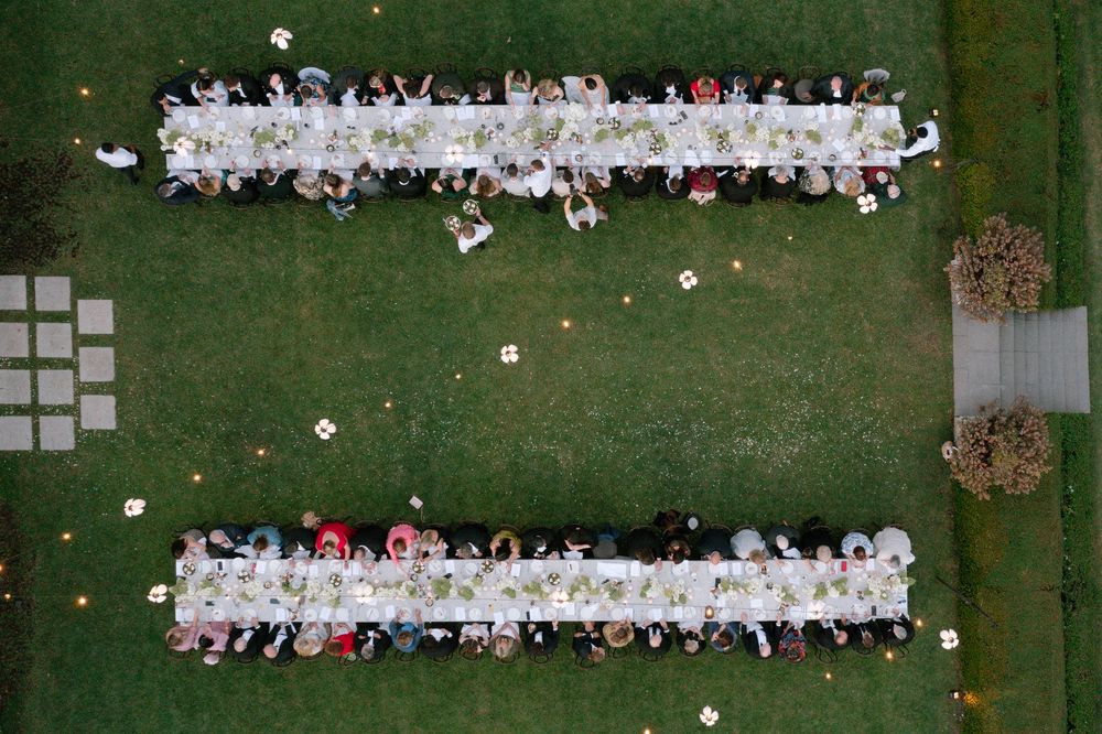 Aerial view of two long elegant outdoor wedding reception tables on green lawn with guests seated on both sides.