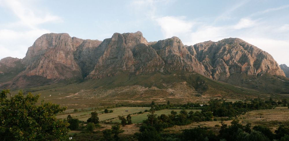Panoramic view of rugged rocky mountain range with green valley and trees under partly cloudy sky.