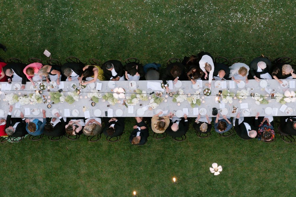 Aerial view of guests seated at a long elegant outdoor dining table on green grass.
