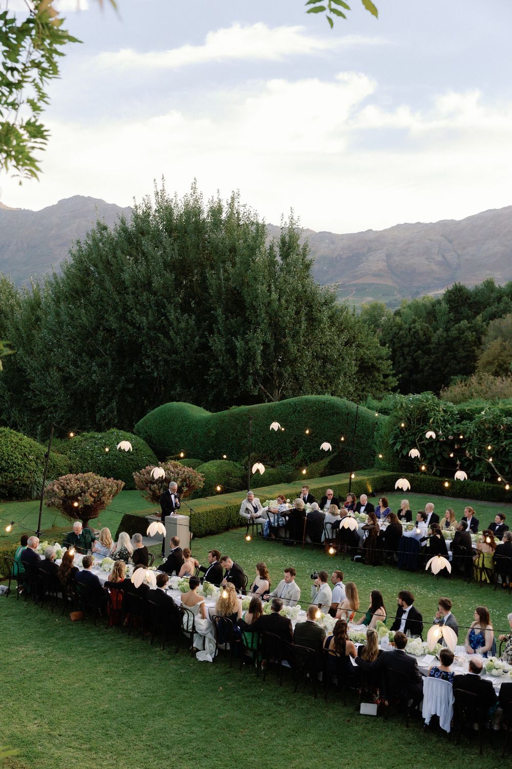 Elegant outdoor wedding reception on lush green lawn with mountain backdrop, string lights, and guests dining at long tables.