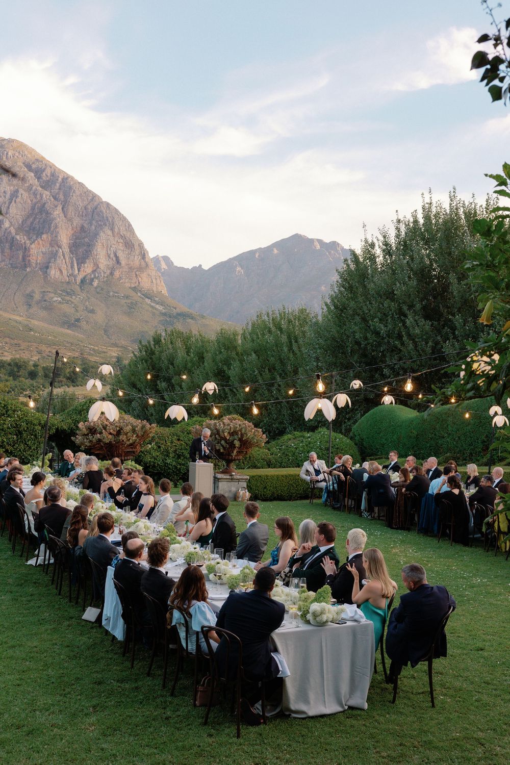 Elegant outdoor wedding reception with string lights, mountain backdrop, and guests seated at long tables on lush green lawn.