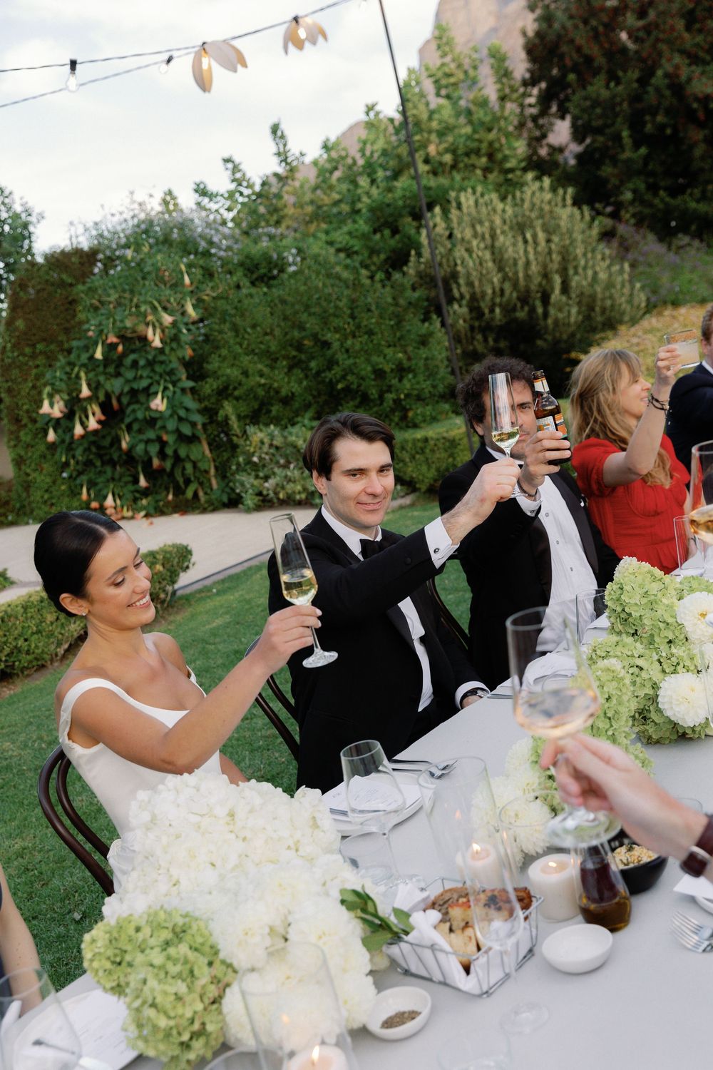 Bride and groom toasting champagne with guests at elegant outdoor garden wedding reception with white floral centerpieces.