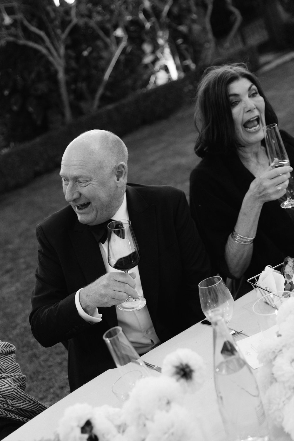 Laughing couple in formal attire raising champagne glasses at an elegant outdoor wedding reception dinner.