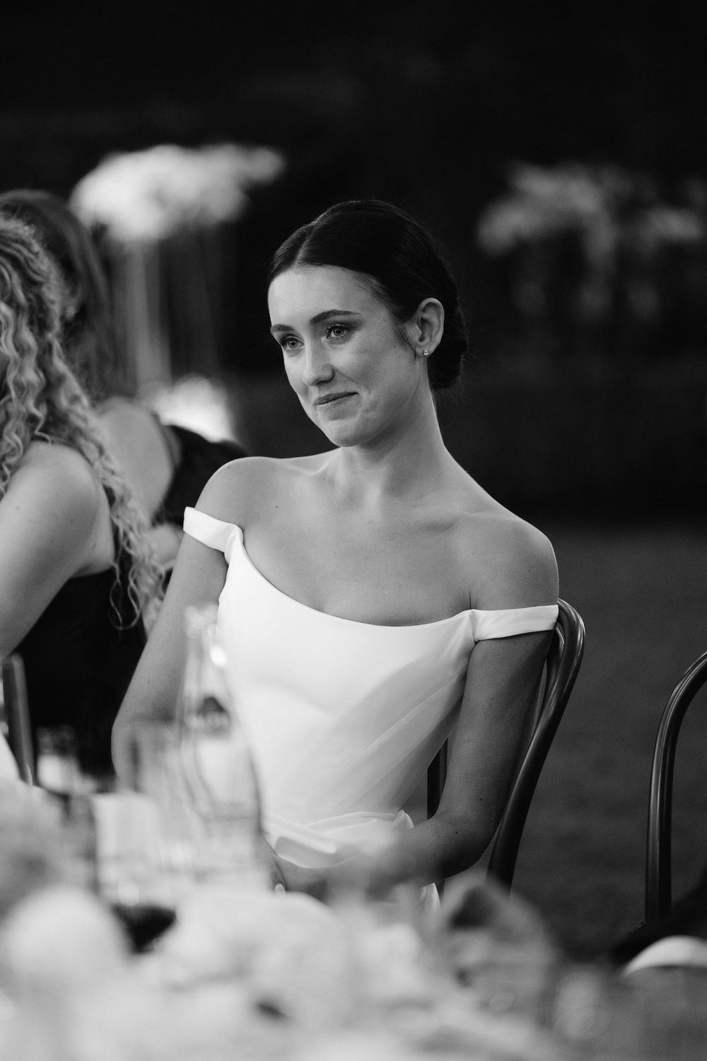 Elegant bride in off-shoulder white gown seated at wedding reception table, black and white photography.
