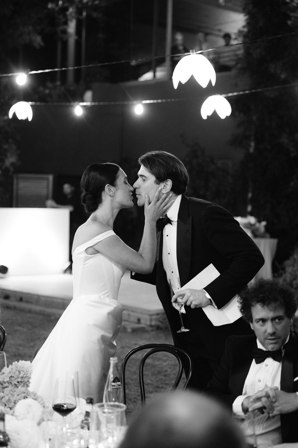 Bride and groom sharing a romantic kiss at their candlelit outdoor wedding reception under string lights.