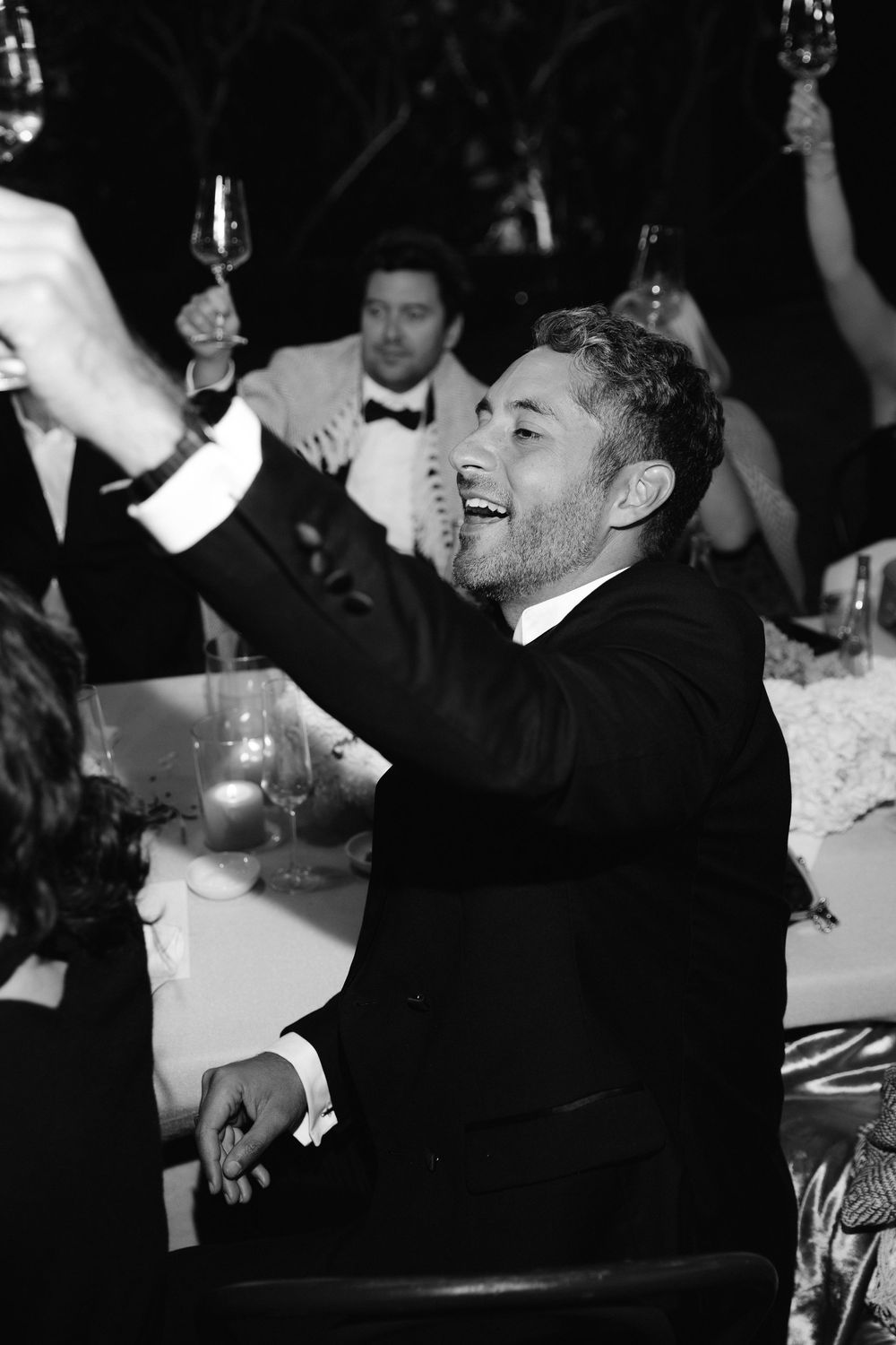 Black and white photo of a man in a tuxedo raising a champagne glass at a formal wedding reception.