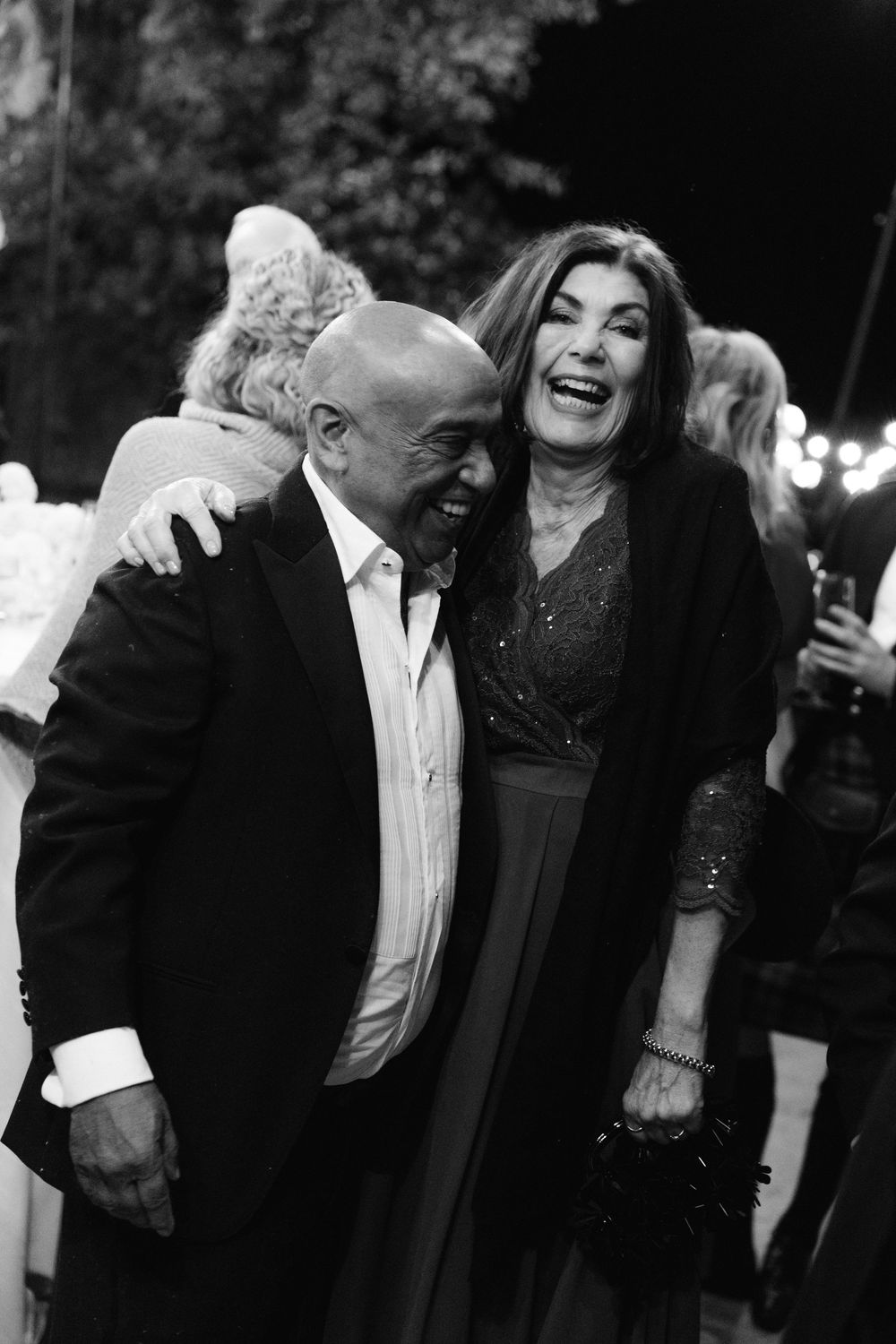 Joyful black and white photo of two wedding guests laughing and embracing at an outdoor evening reception.