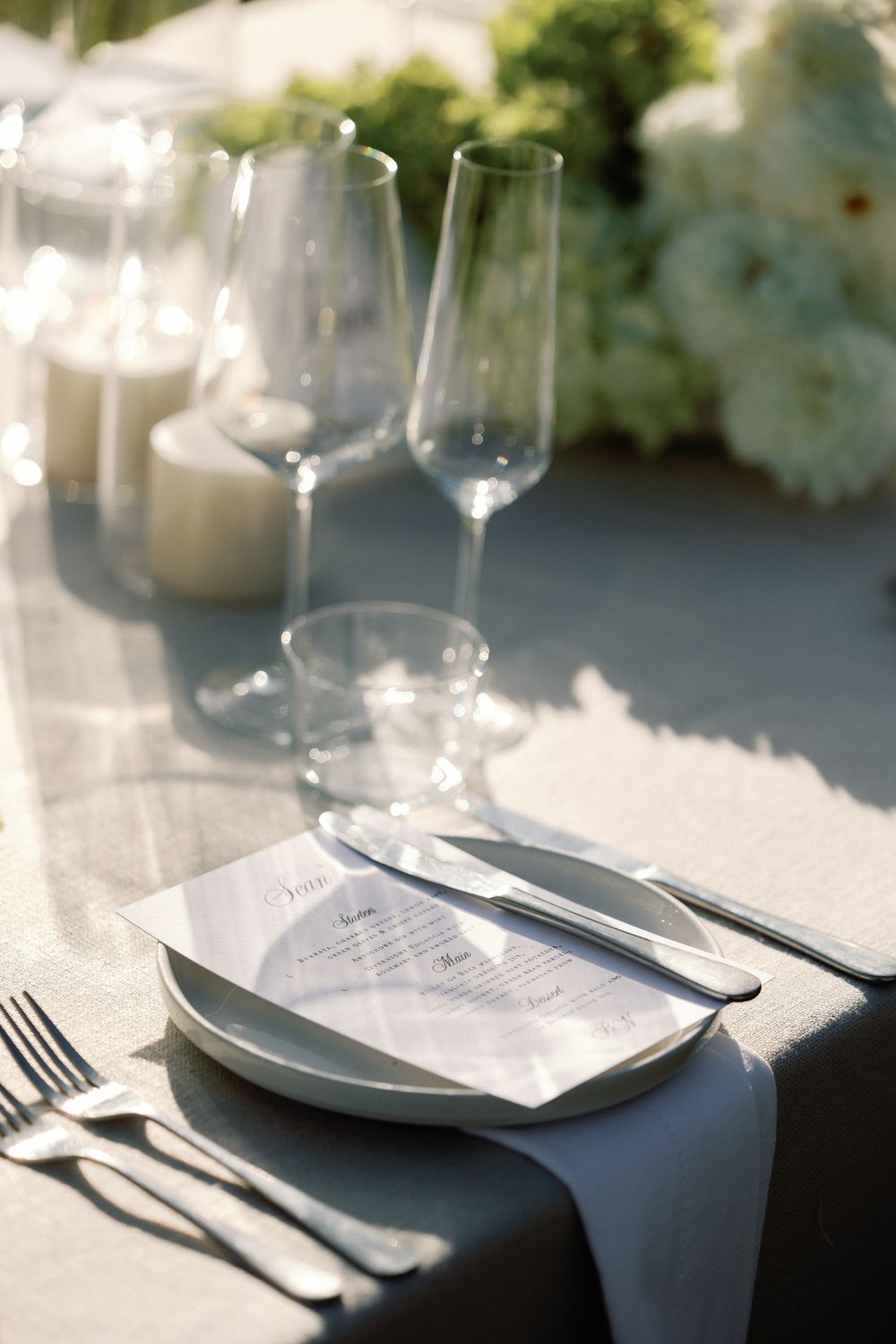 Sophisticated wedding place setting with white menu card, crystal stemware, silver cutlery, and gray linen napkin.