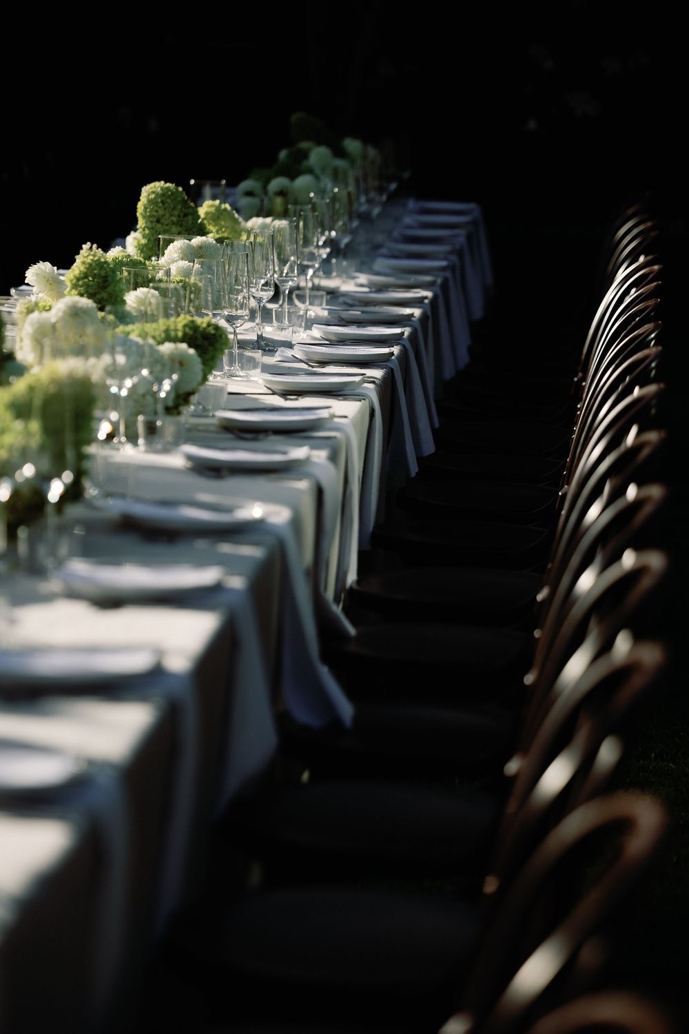 Elegant long banquet table set with white linens, crystal glasses, and lush green floral centerpieces in a dimly lit venue.