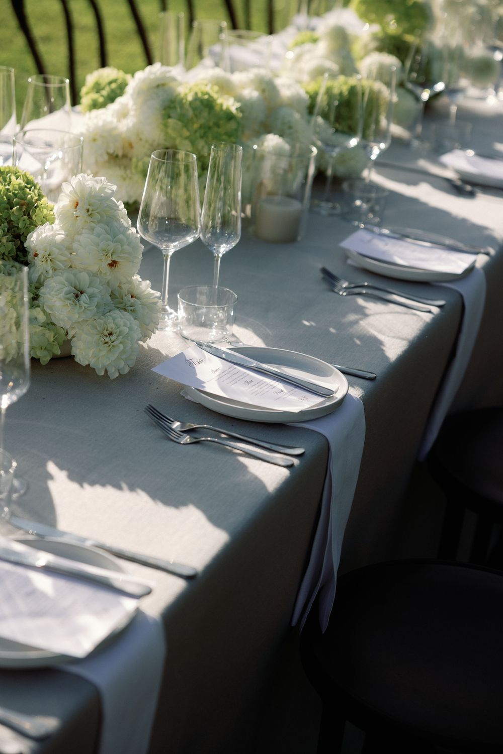 Elegant wedding reception table set with white floral centerpieces, crystal glasses, and silver cutlery on a gray linen tablecloth.
