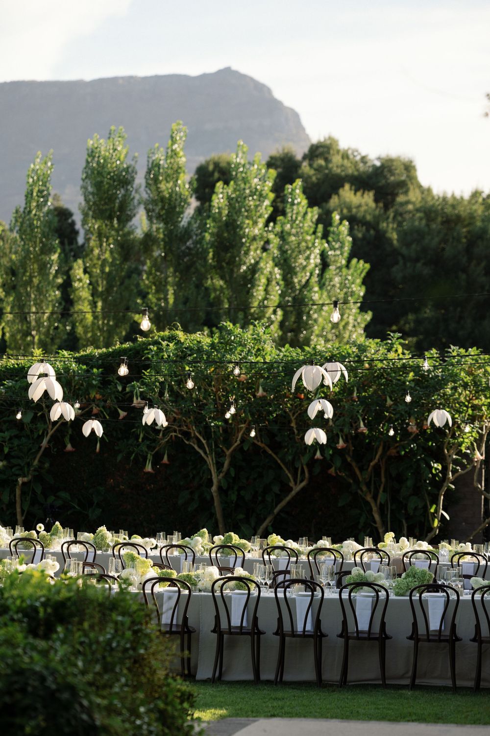 Outdoor garden wedding reception with long white tables, bistro chairs, hanging floral decor, and mountain backdrop.