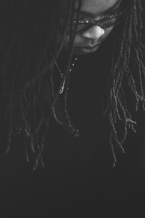 Black and white portrait of a person with long dreadlocks, wearing sunglasses and a necklace, looking downward.