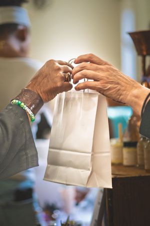 Two people exchanging a white paper takeout bag over a counter in a restaurant or cafe setting.