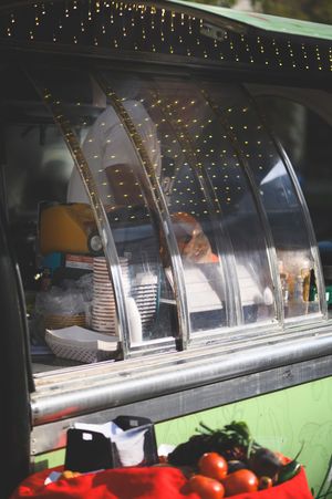 A food truck vendor behind curved glass display with fresh tomatoes and vegetables on the counter outside.