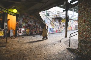 Colorful graffiti-covered walls line a cobblestone alley with a costumed bear character near a coffee shop entrance.