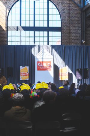 Crowd wearing yellow hard hats at a BUILD Detroit event inside a brick building with large arched windows.