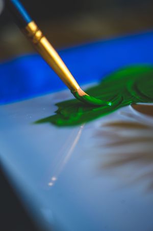 A fine-tipped paintbrush with gold ferrule applying green paint onto a canvas in a close-up artistic shot.