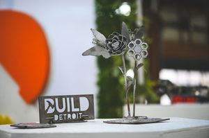 Metal sculpture of a rose and flower beside a Build Detroit sign on a white table at an event.