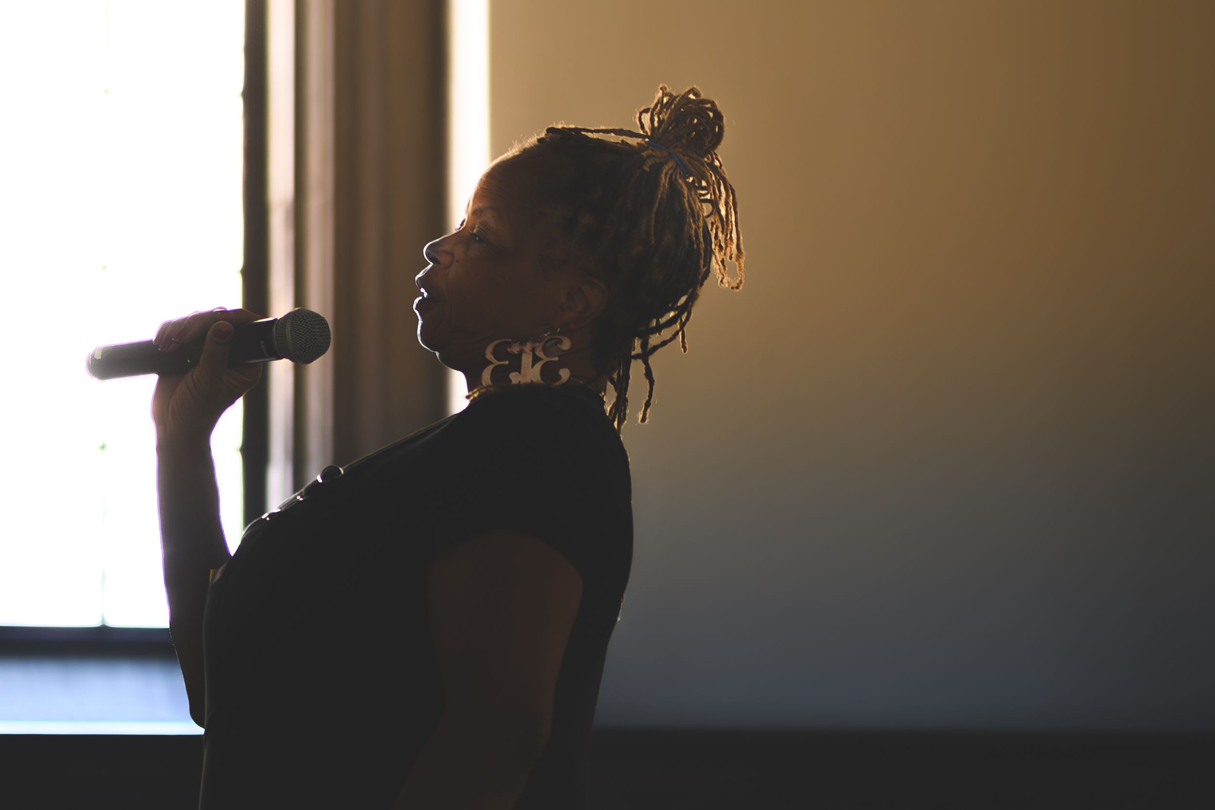 Silhouette of a person with braided hair holding a microphone, backlit by a window in a dramatic performance setting.