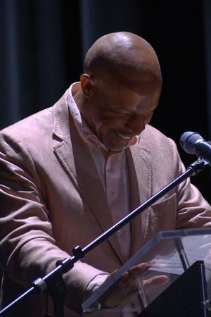 Smiling bald man in beige blazer speaking at a clear podium with a microphone at a formal event.