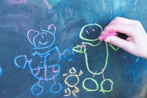 Child's hand drawing colorful chalk figures of people on a dark chalkboard surface.